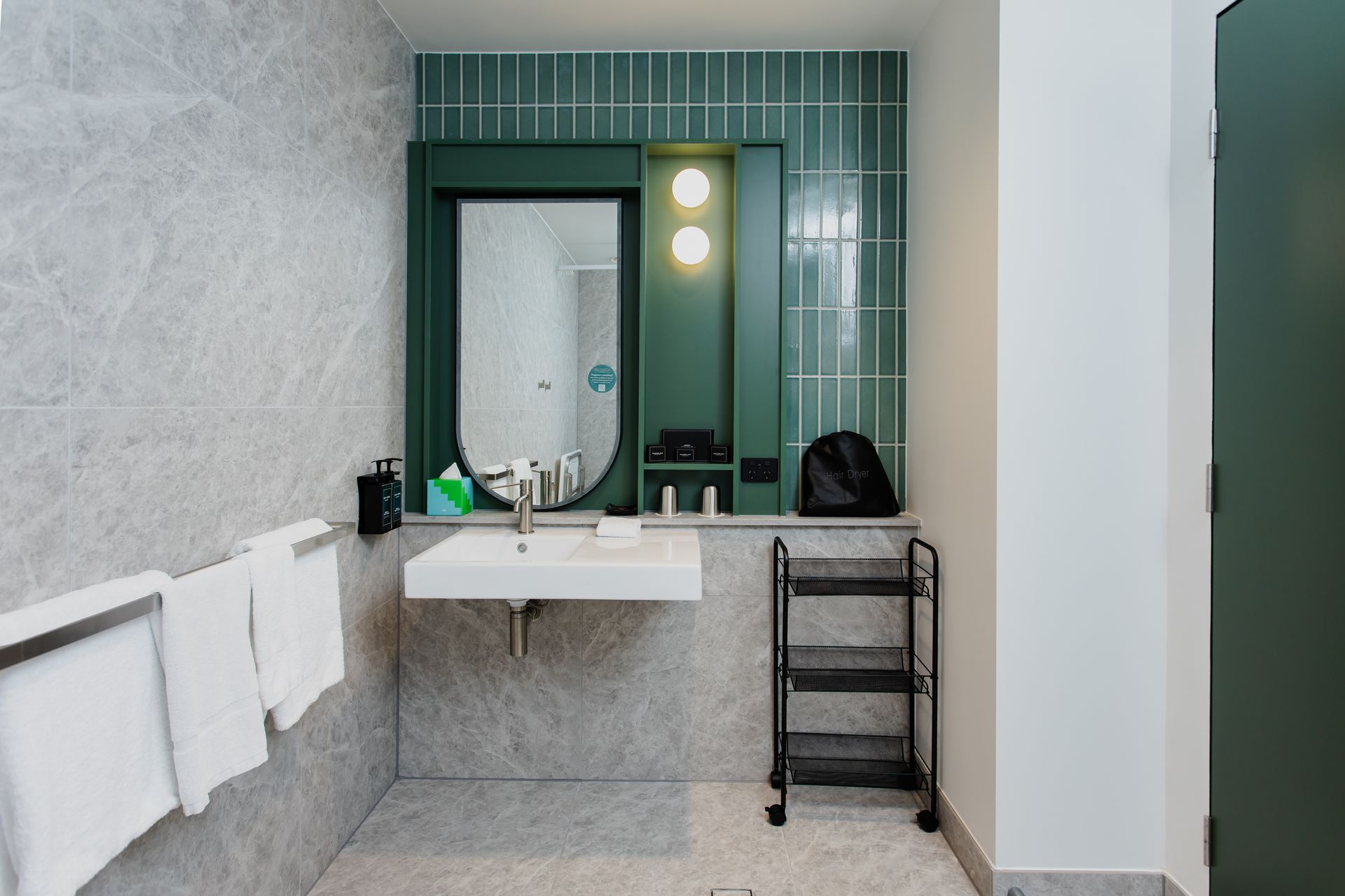 Modern bathroom with gray tiled walls and a green-framed mirror, white sink, and black rolling cart.