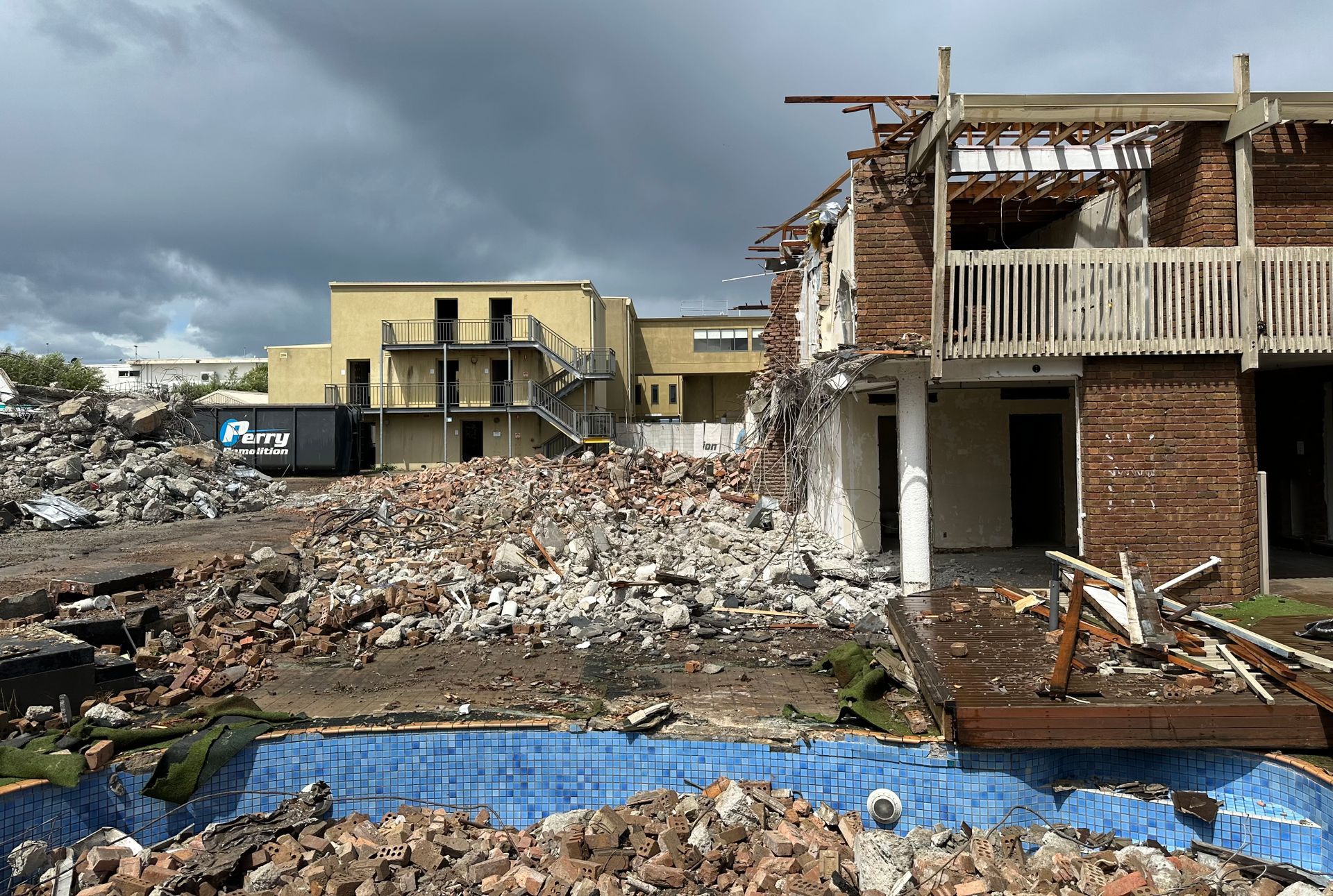 Demolished brick building ruins next to a drained pool and a yellow building under an overcast sky.