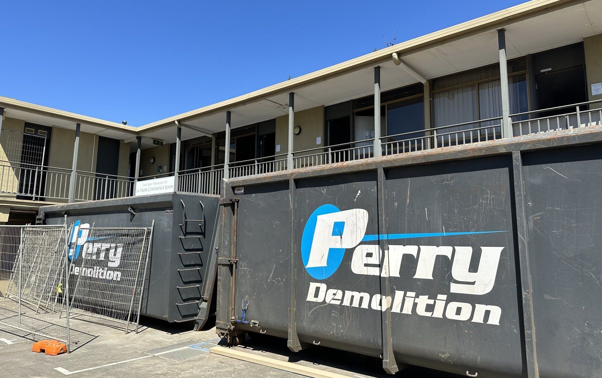 Two dark dumpsters from Perry Demolition are parked near a beige building. A chain link fence is in the foreground.