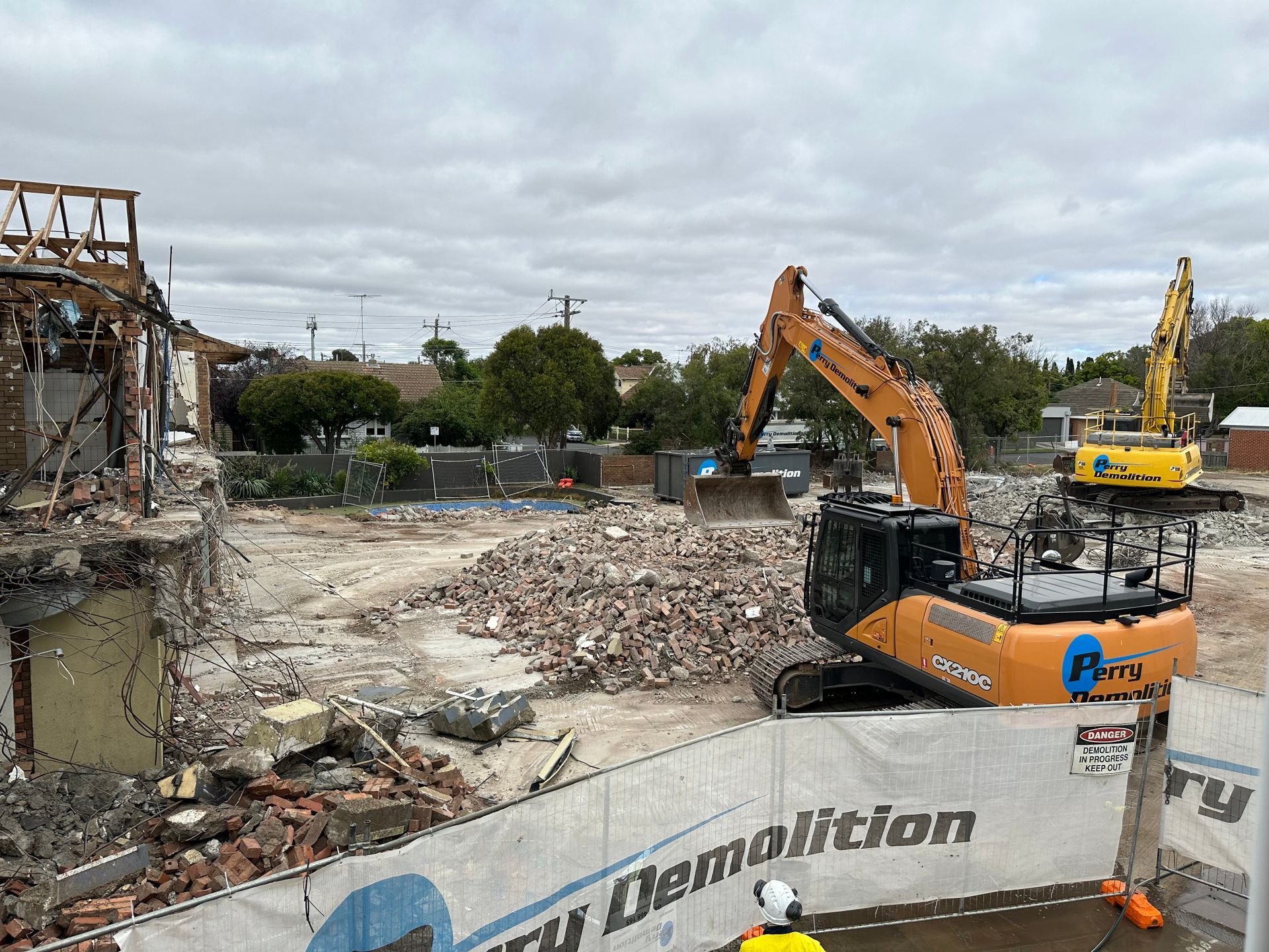 Demolition site with orange and yellow excavators, debris, and a partially demolished building on a cloudy day.