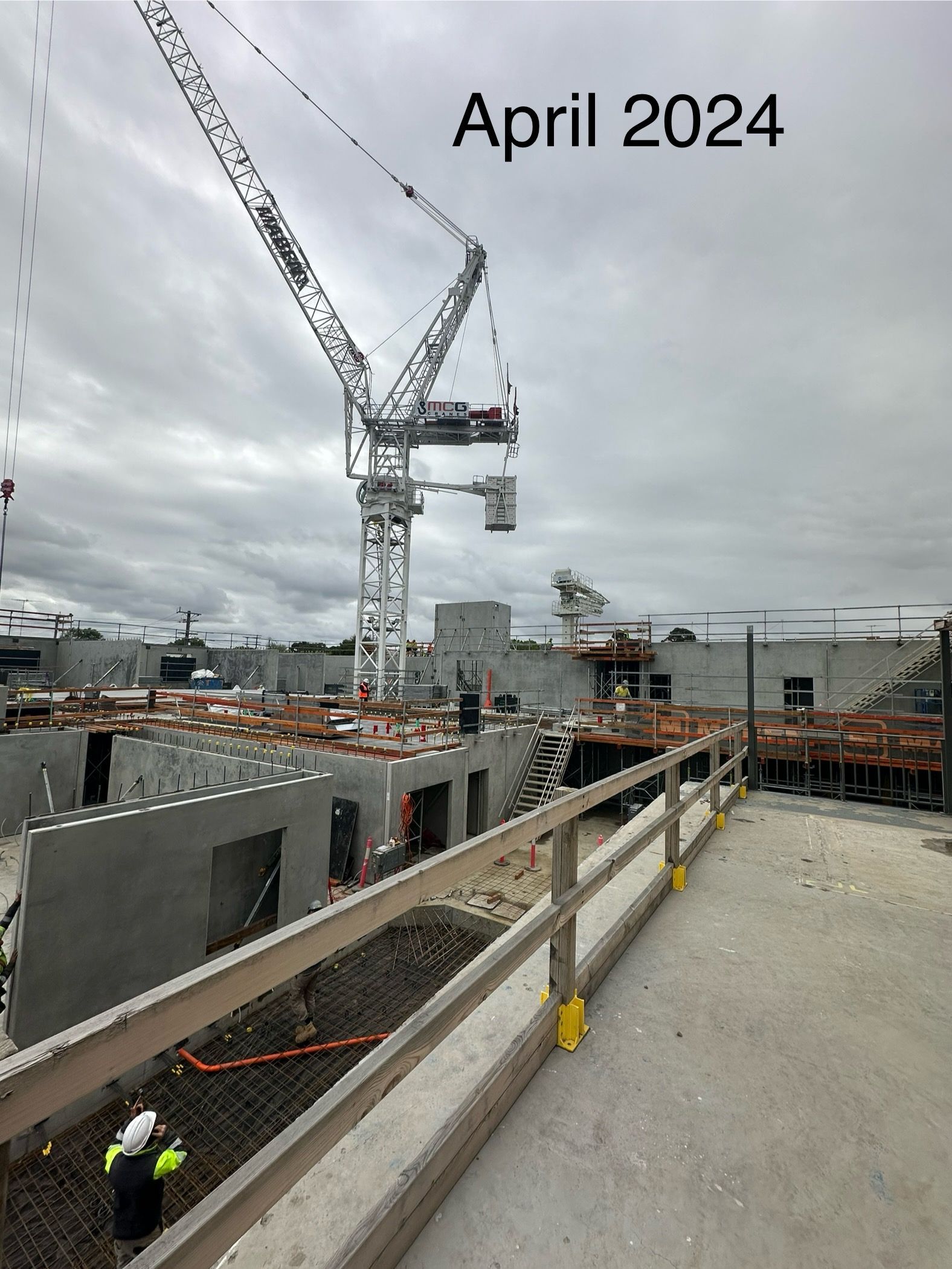 Construction site in April 2024 with tower cranes, partially built concrete structures, and safety barriers.
