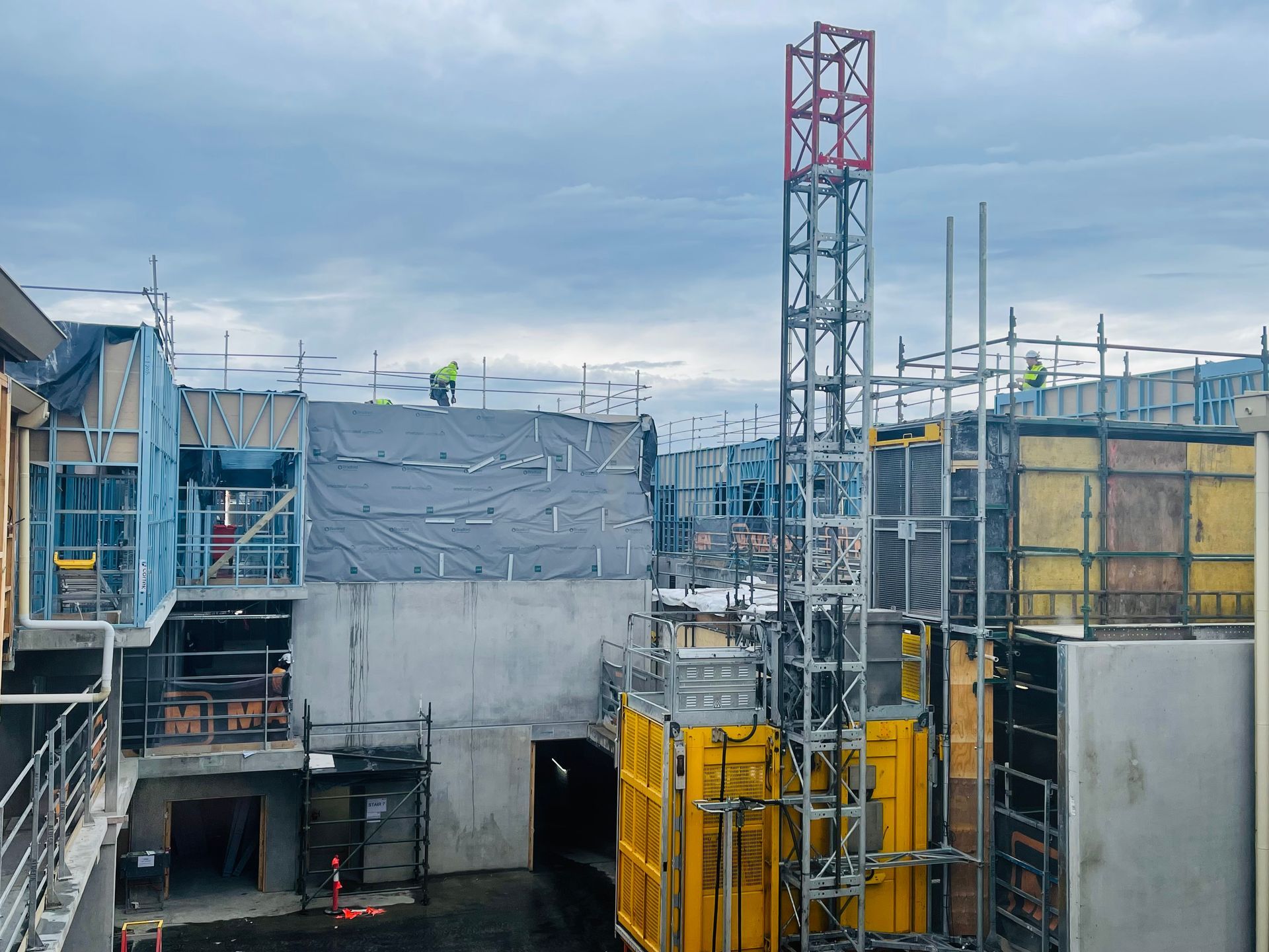 Construction site with a yellow hoist, gray concrete, and blue scaffolding under an overcast sky.