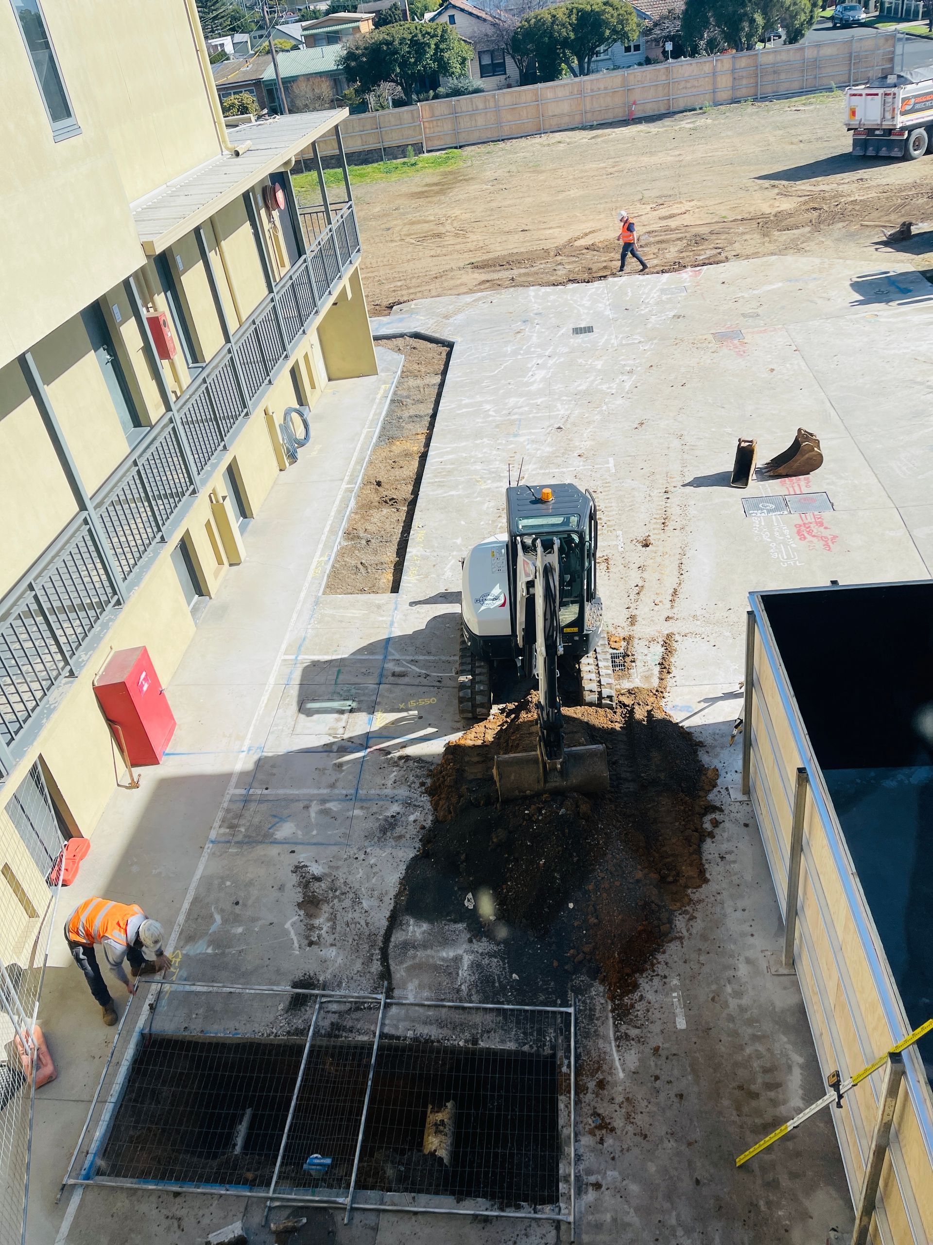 Construction site: excavator digging on concrete near a building; workers in safety vests.