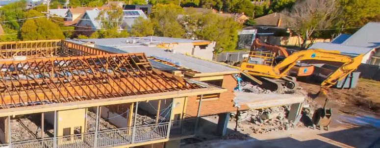 An excavator demolishes a building; rooftops and trees in the background.