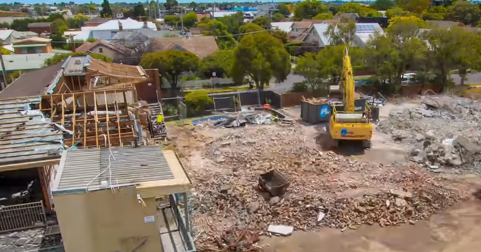 Construction site with excavator demolishing a building, surrounded by debris and houses.