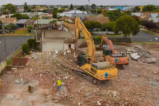 Demolition site with two excavators tearing down a building, debris scattered on the ground, and a worker.