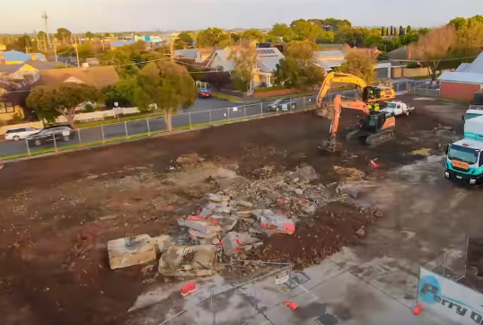 Construction site with an excavator, cleared dirt, and surrounding buildings.