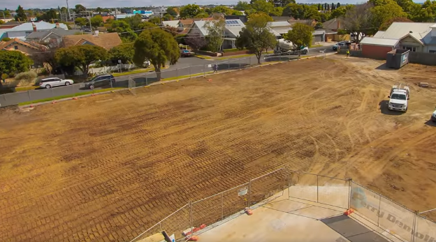 Cleared construction site with dirt, surrounded by a fence, houses, and parked vehicles.