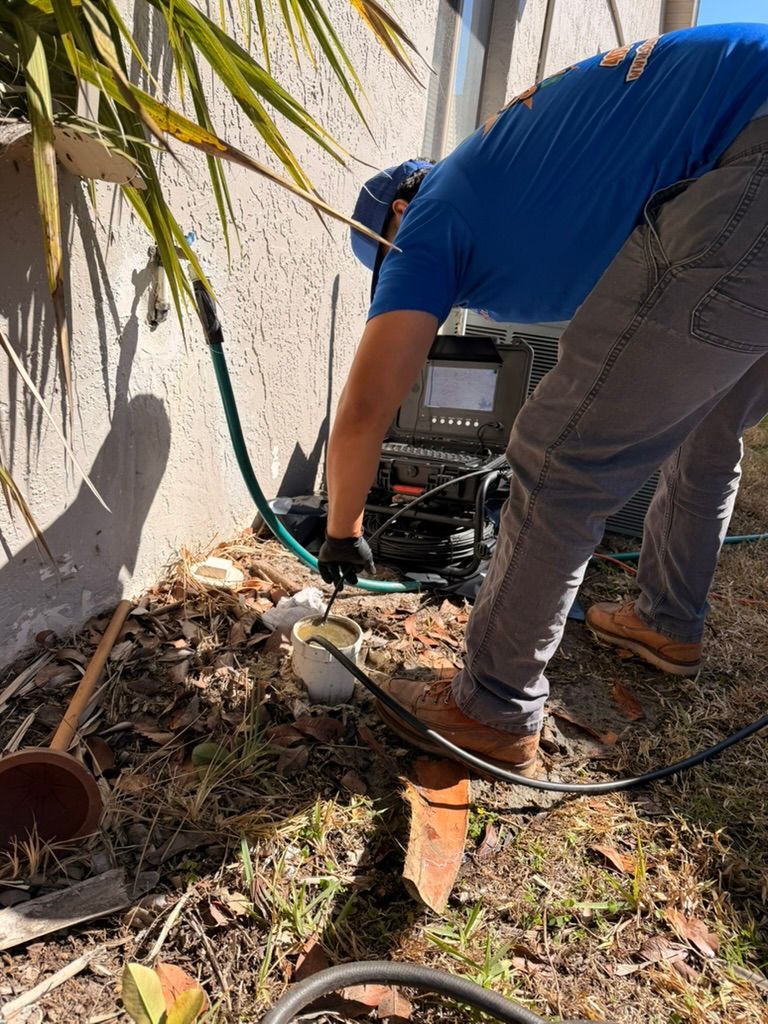 A person in a blue shirt uses a drain inspection camera system to examine a ground-level pipe outside a house.