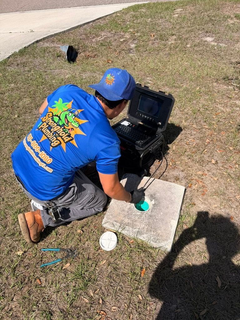 A technician in a blue shirt uses a pipe inspection camera to check an underground sewer line in a grass yard.
