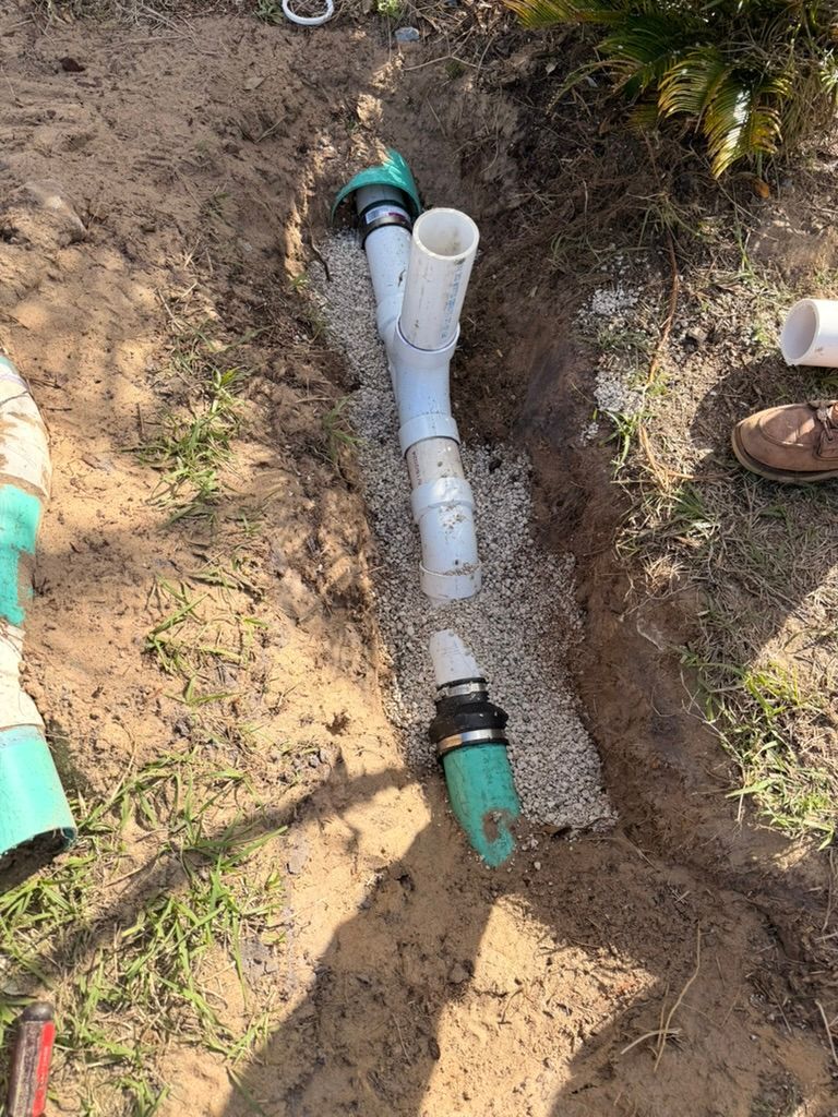 White PVC pipe Y-connector installed in a narrow dirt trench filled with gravel, connecting green drainage pipes.