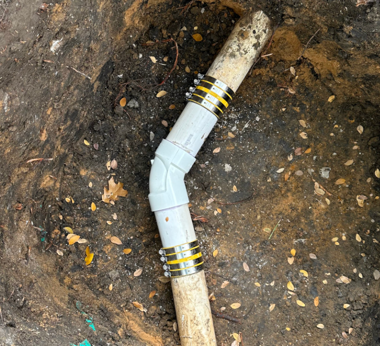 A white PVC pipe angled fitting connected to two older pipes in the dirt using two rubberized metal sleeve couplings.