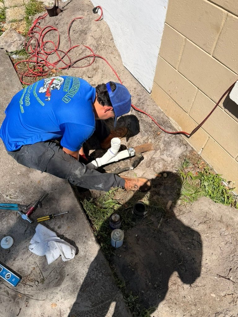 A plumber in a blue uniform kneels outside near a building wall, working on an exposed white PVC pipe in the ground.