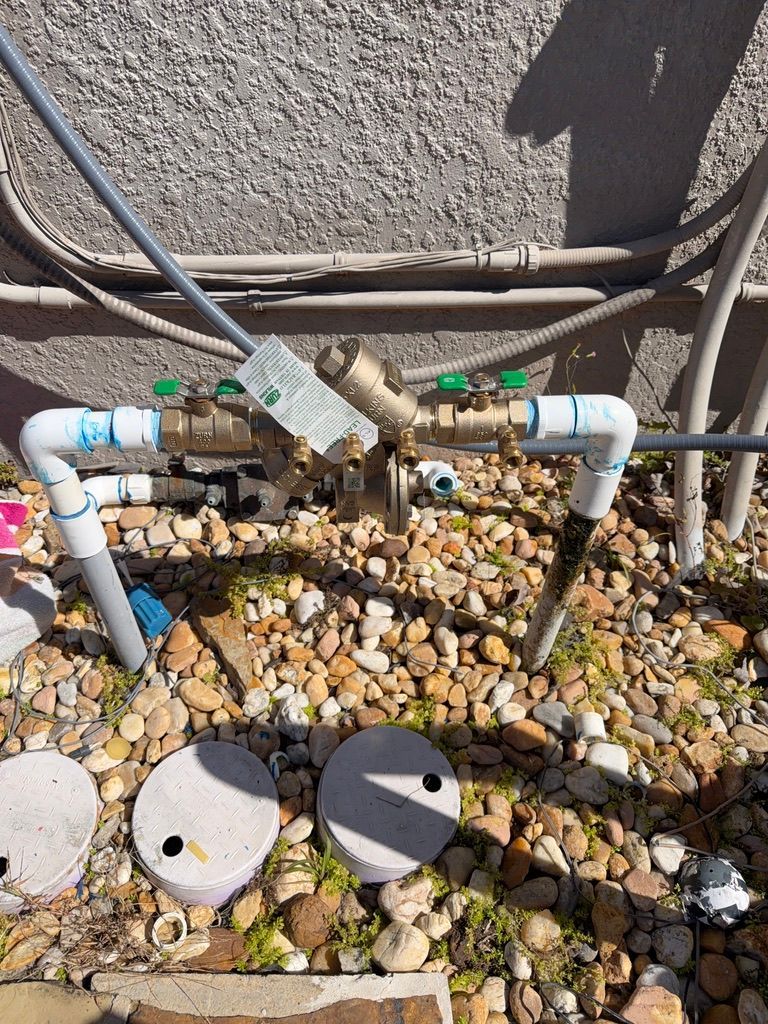 An outdoor brass backflow prevention assembly connected to white PVC pipes, installed on gravel ground by a house wall.