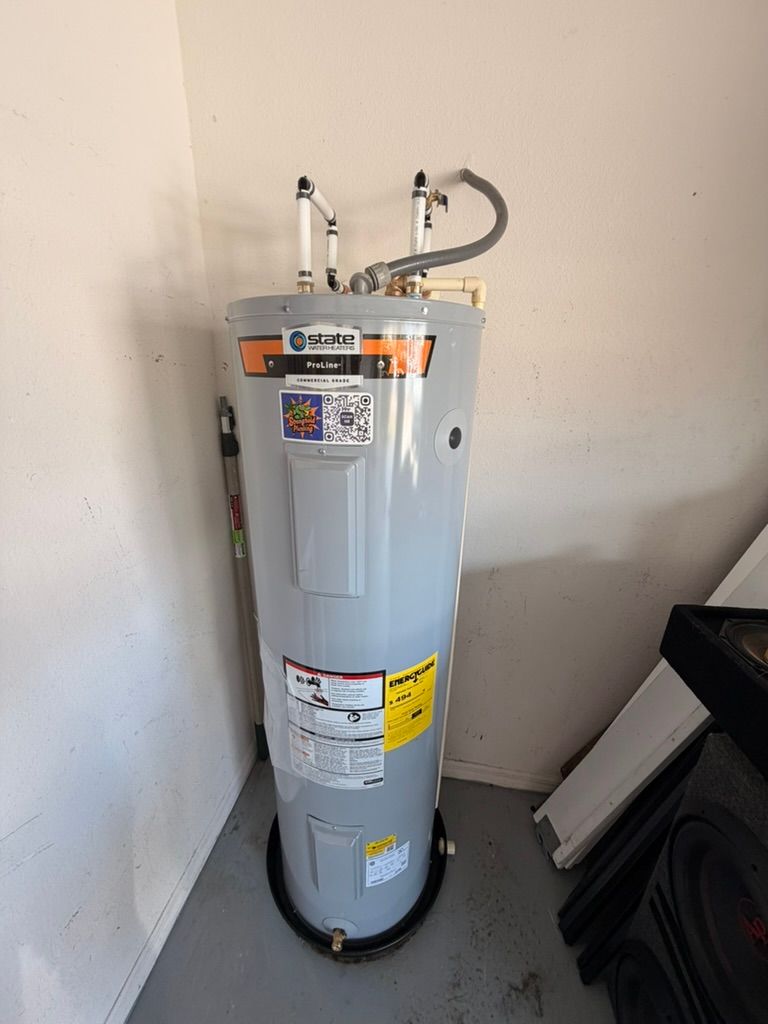 A gray residential electric water heater stands in the corner of a room, with white plumbing pipes connected to the top.