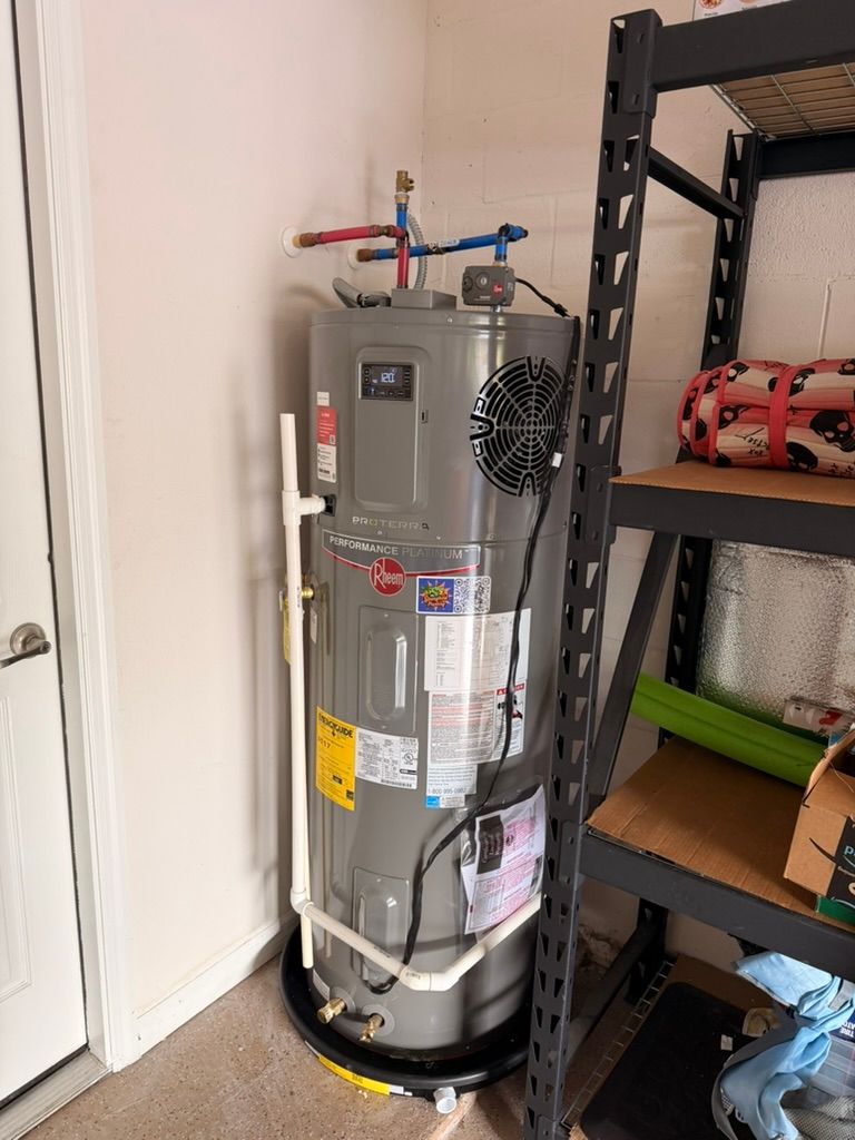 A gray Rheem hybrid electric water heater standing in a drain pan next to a black metal storage shelf in a garage.