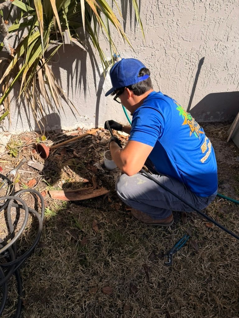 A person wearing a blue shirt and hat crouches outside to inspect a water spigot near a stucco wall and palm plant.