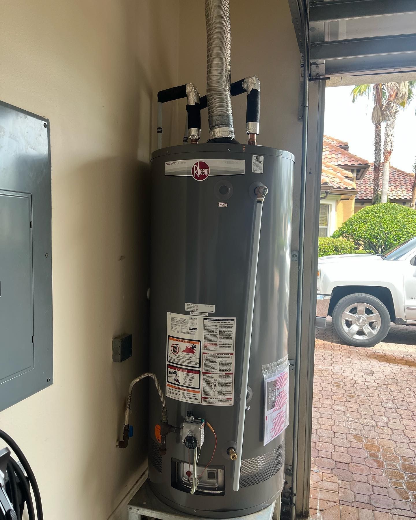 A tall, gray, cylindrical gas water heater stands in a garage next to a gray electrical panel and an open doorway.
