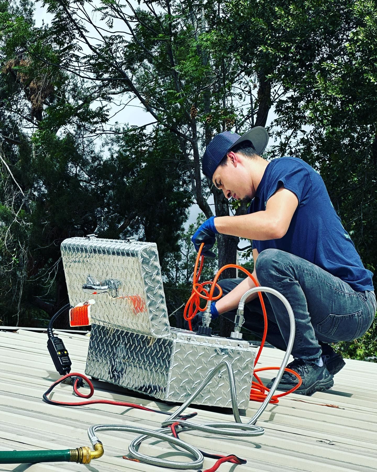 A person in a blue shirt and cap works with tools and cables from an open metal toolbox on a corrugated roof.