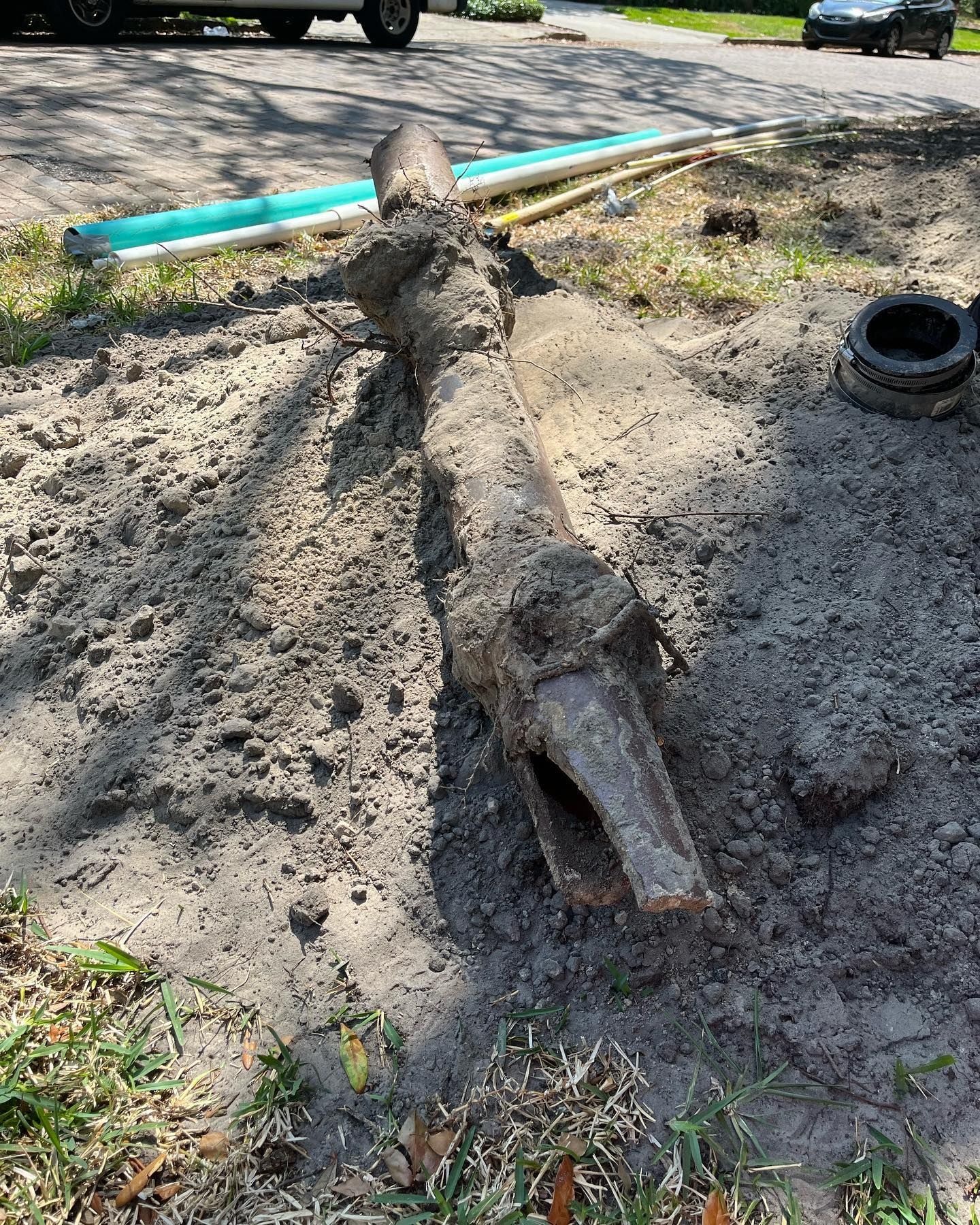 A long, dirt-covered metal pipe section lies in the soil near a road, with new pipe segments visible in the background.