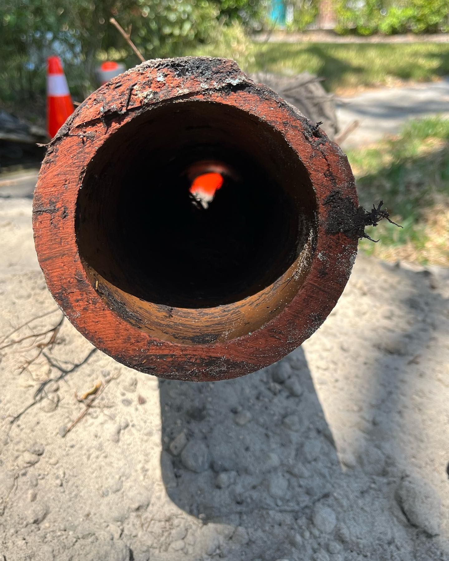 A close-up view through a thick, rust-colored circular pipe, revealing a small orange traffic cone in the distance.