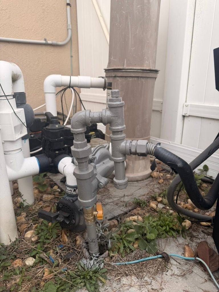 Outdoor pool plumbing setup with white PVC pipes and gray metal fittings, positioned near a fence and pump equipment.