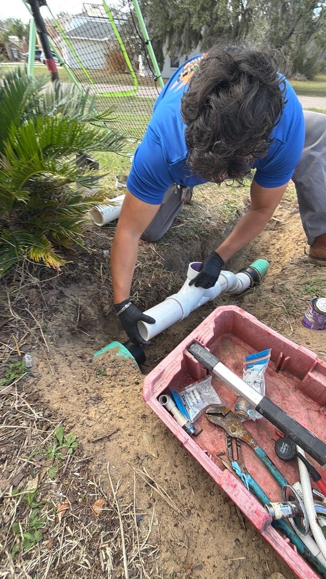 A person wearing a blue shirt and black gloves connects white PVC pipe in a dirt trench next to a container of tools.
