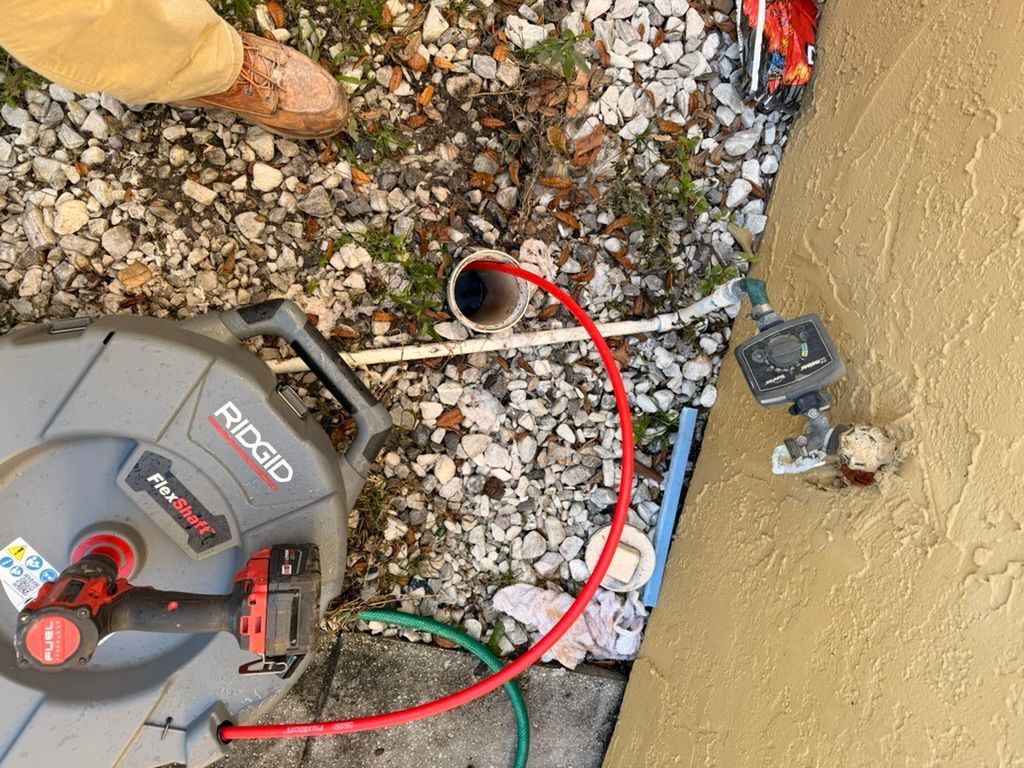 A RIDGID camera reel sits on gravel next to a drainpipe and a home exterior wall with a water spigot and timer.