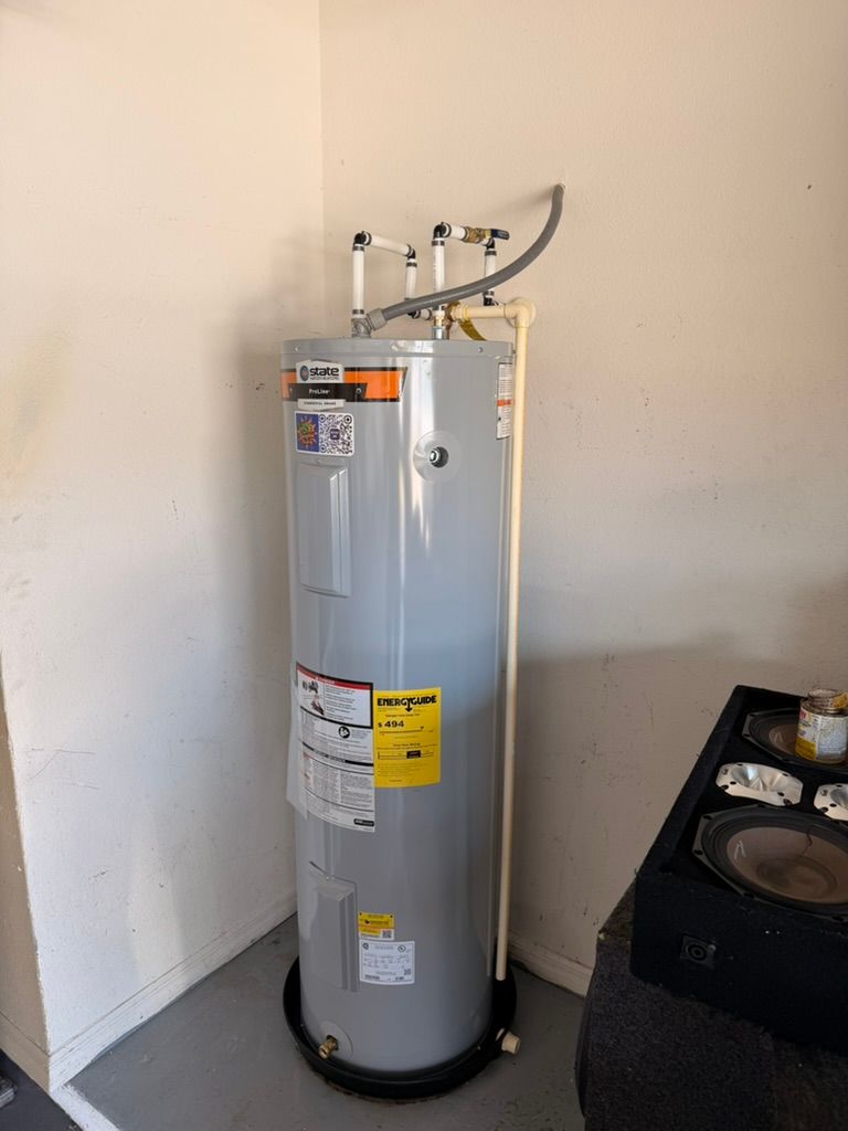 A tall, grey electric water heater stands in a corner on a black drain pan, connected by white pipes to the wall.