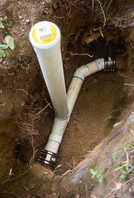 A white PVC sewer cleanout pipe installed in a trench, connected to existing underground piping with rubber couplings.