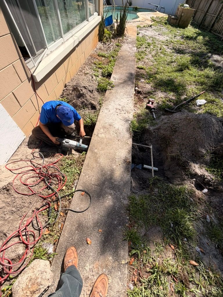 A person in a blue shirt uses a power tool to cut into the dirt near a concrete walkway and house exterior.