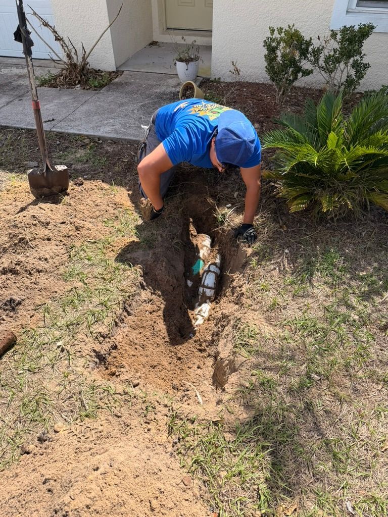A person in a blue shirt kneels in a yard, digging into the soil with their hands to access buried white PVC piping.