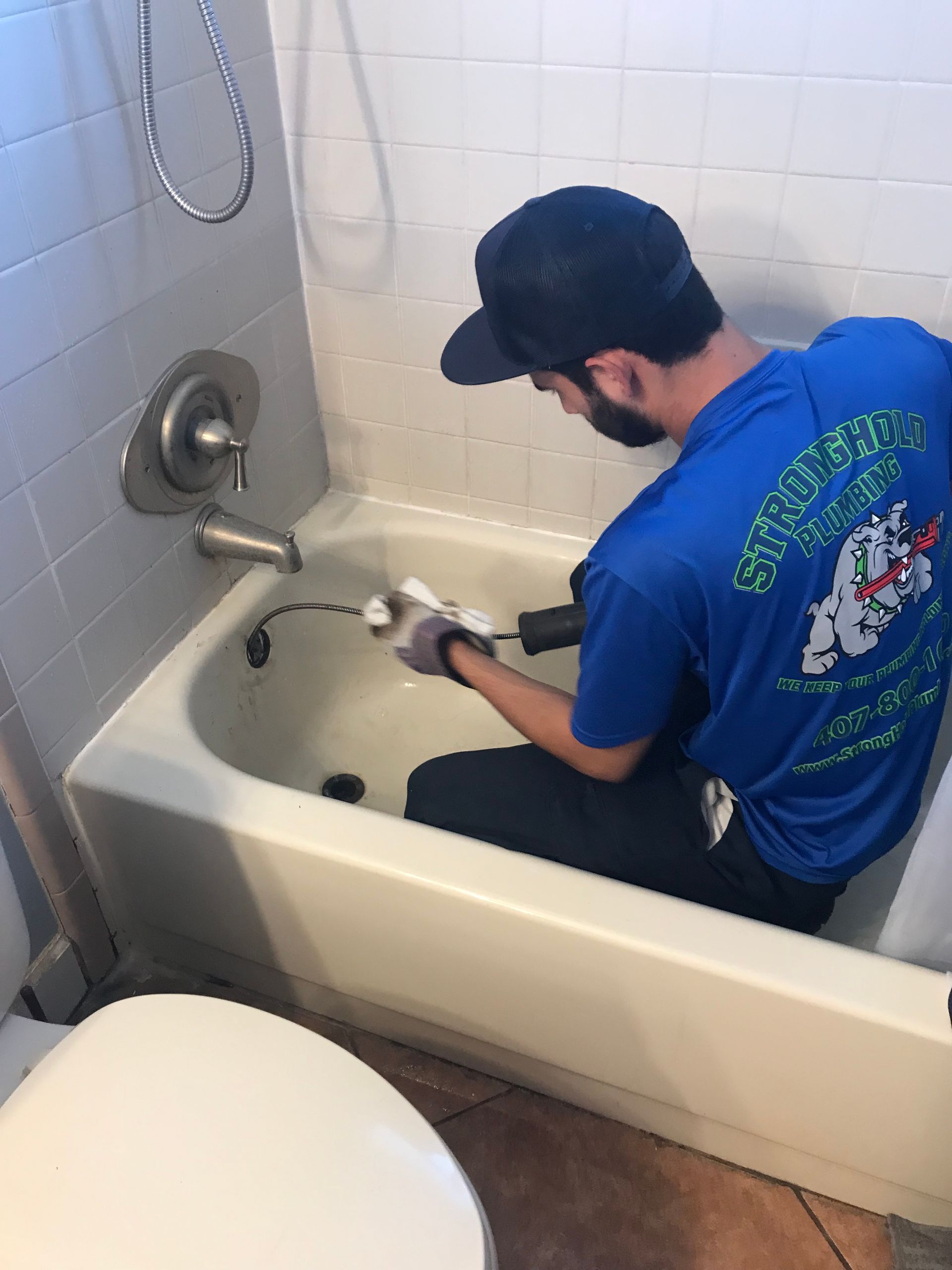 A person wearing a blue uniform shirt and cap works inside a bathtub, using a plumbing tool to clear a drain.