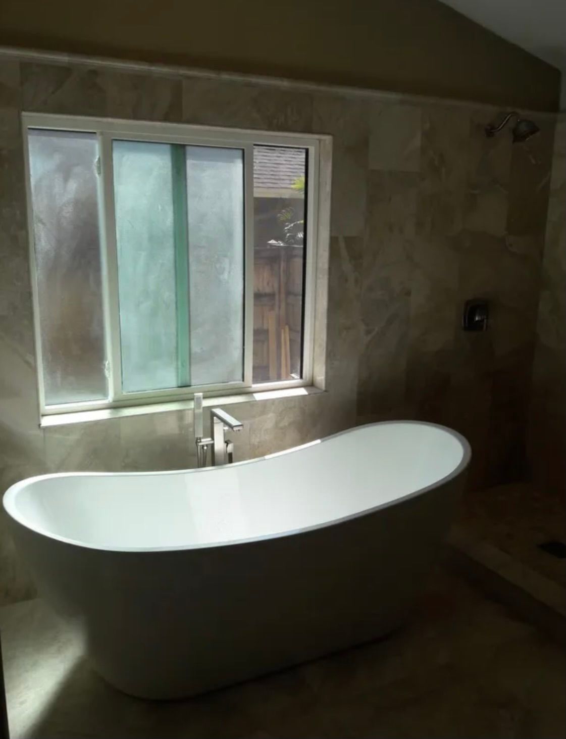 A white freestanding oval bathtub sits in front of a window in a bathroom with tan, stone-tiled walls.