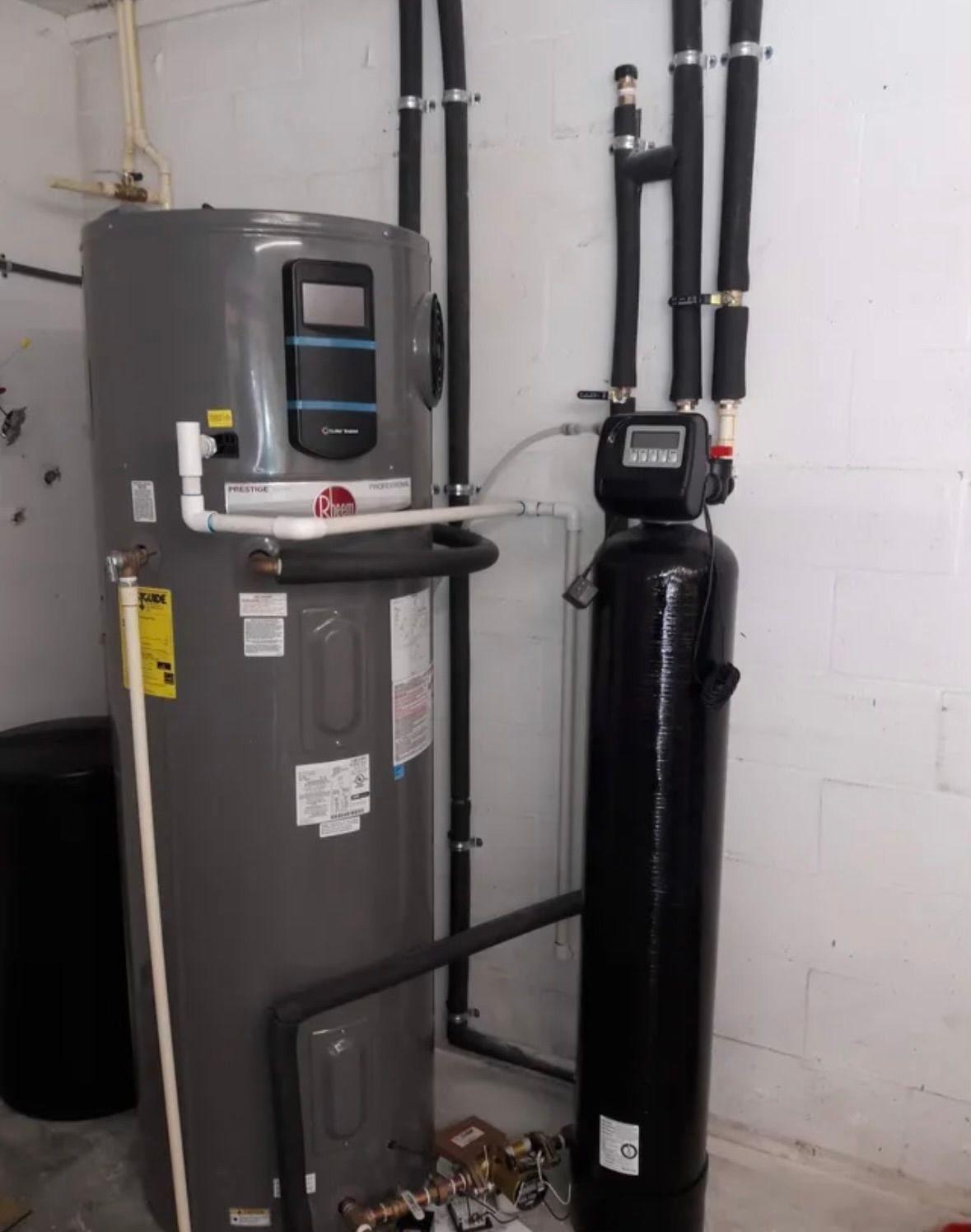 A grey Rheem water heater and a tall black water treatment tank stand against a white wall with insulated piping above.