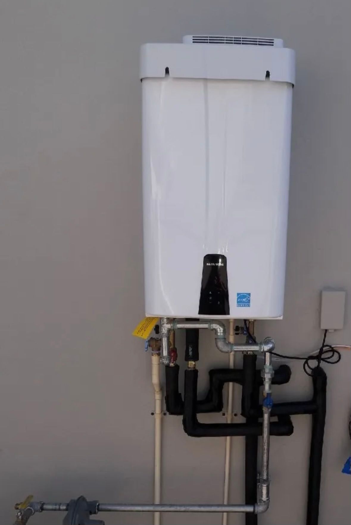 A wall-mounted white tankless water heater with exposed black insulated plumbing pipes against a plain grey wall.