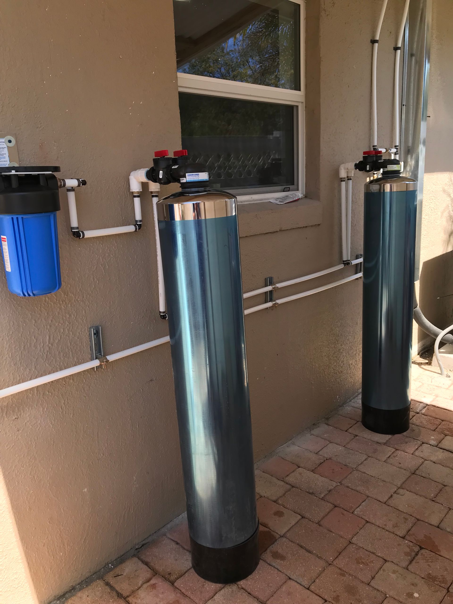 A blue sediment filter and two large grey water treatment tanks installed on the exterior wall of a house.