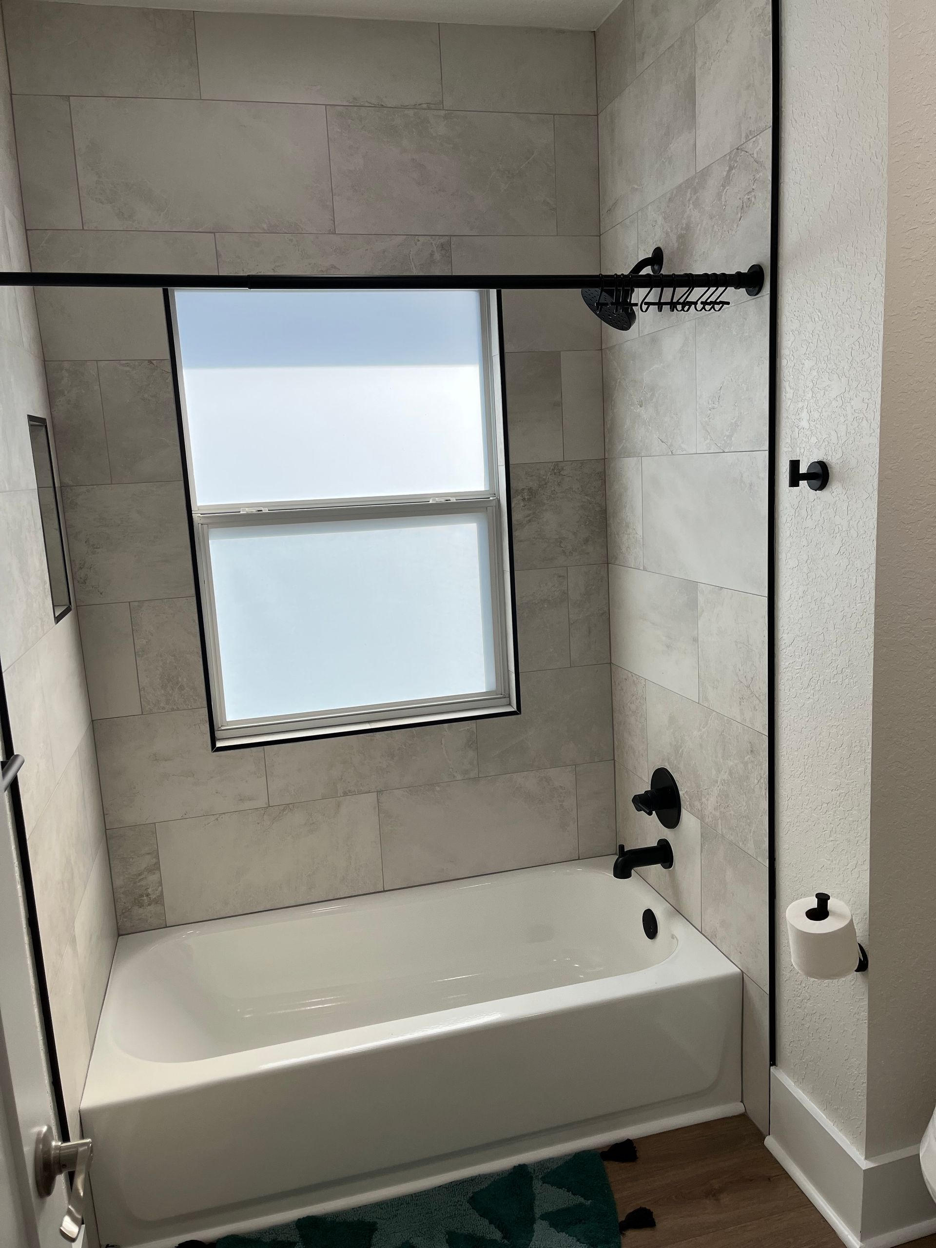 A bathroom shower area featuring light stone-textured wall tiles, a white bathtub, a window, and matte black fixtures.