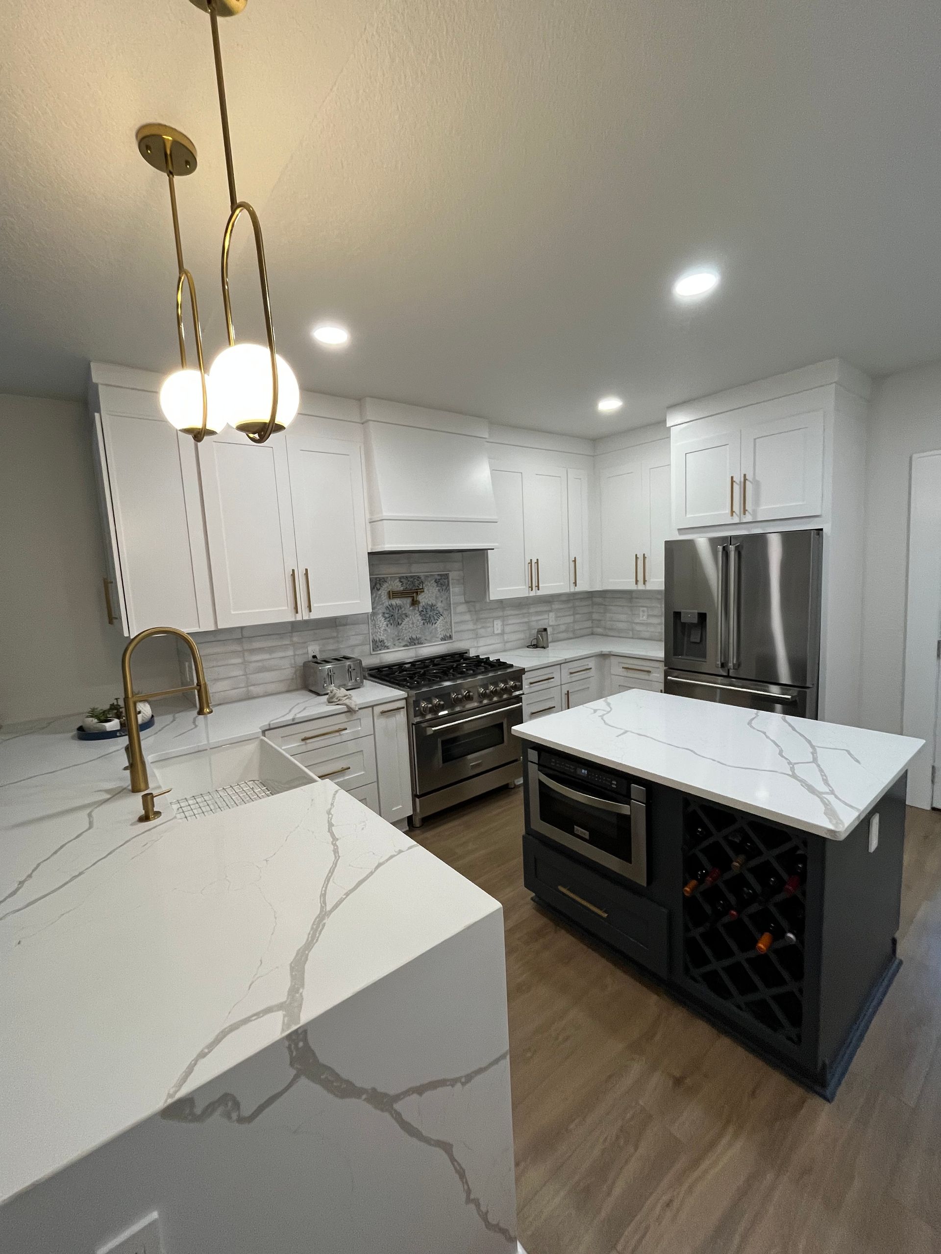 Modern kitchen with a dark-toned island, white cabinets, gold hardware, marble-patterned countertops, and hanging lights.