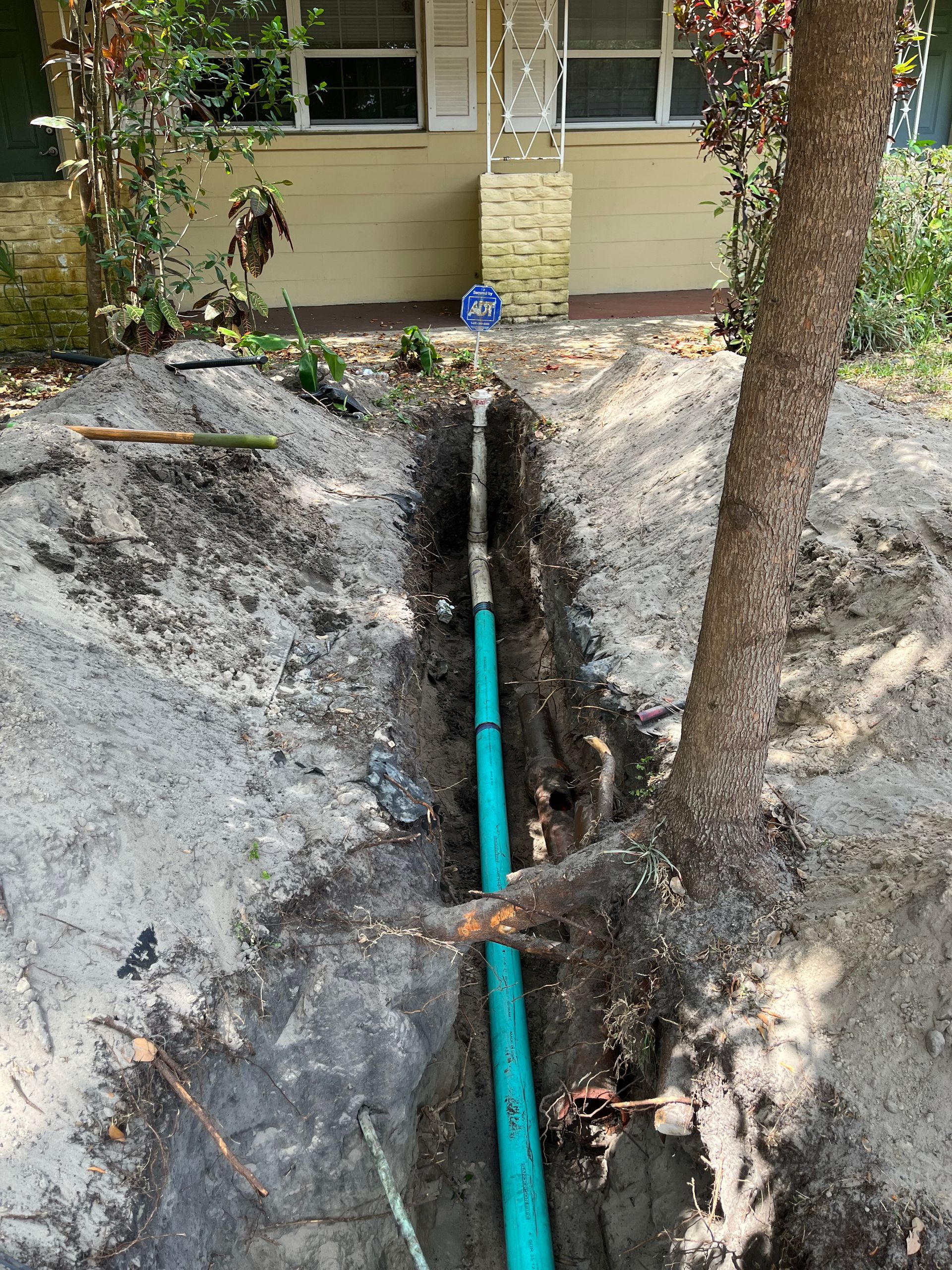 A trench is dug in front of a house, revealing a green pipe running through the center of the dirt path.
