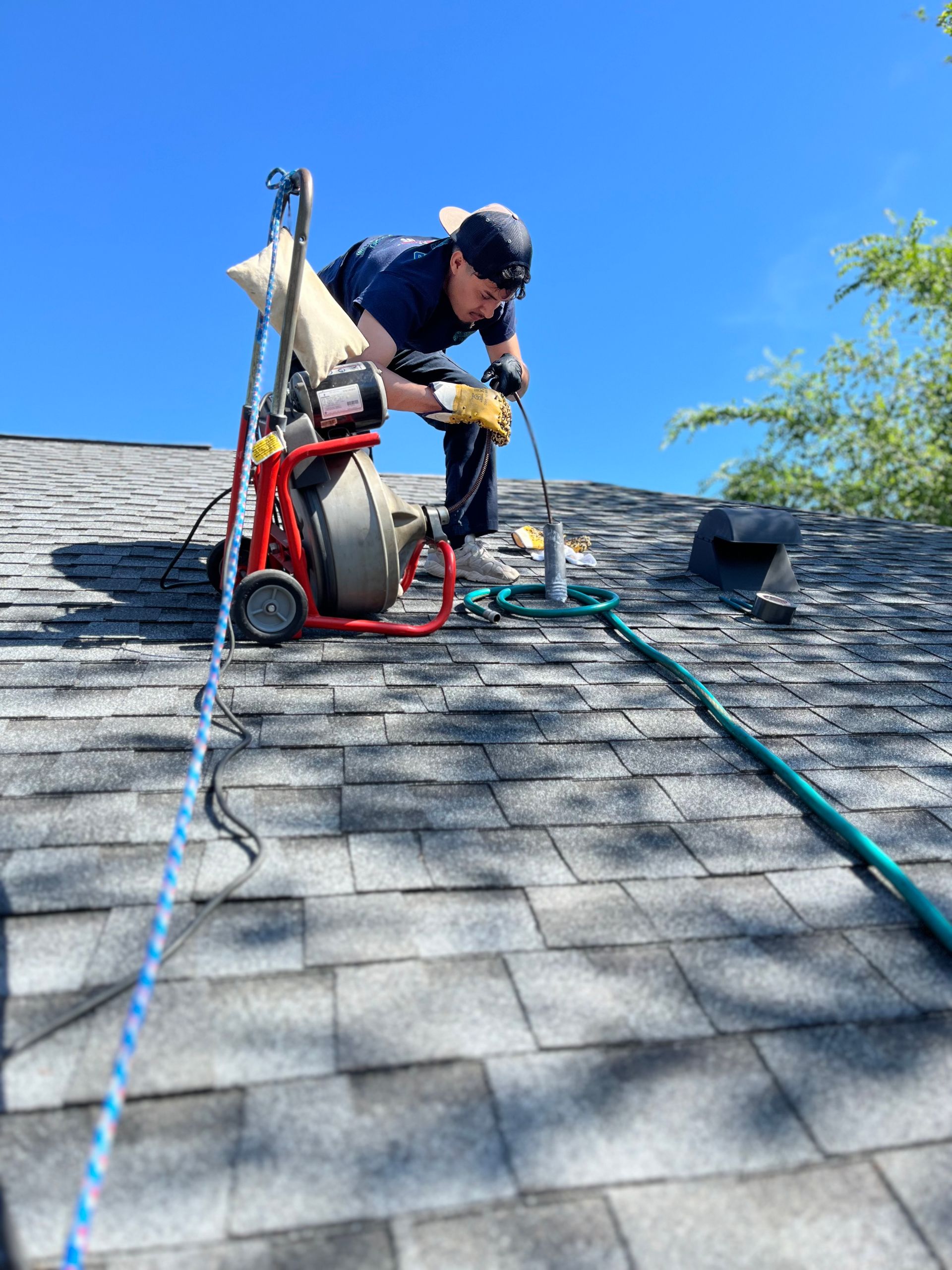 A person wearing a hat and gloves operates a drain cleaning machine on a shingled roof under a clear blue sky.