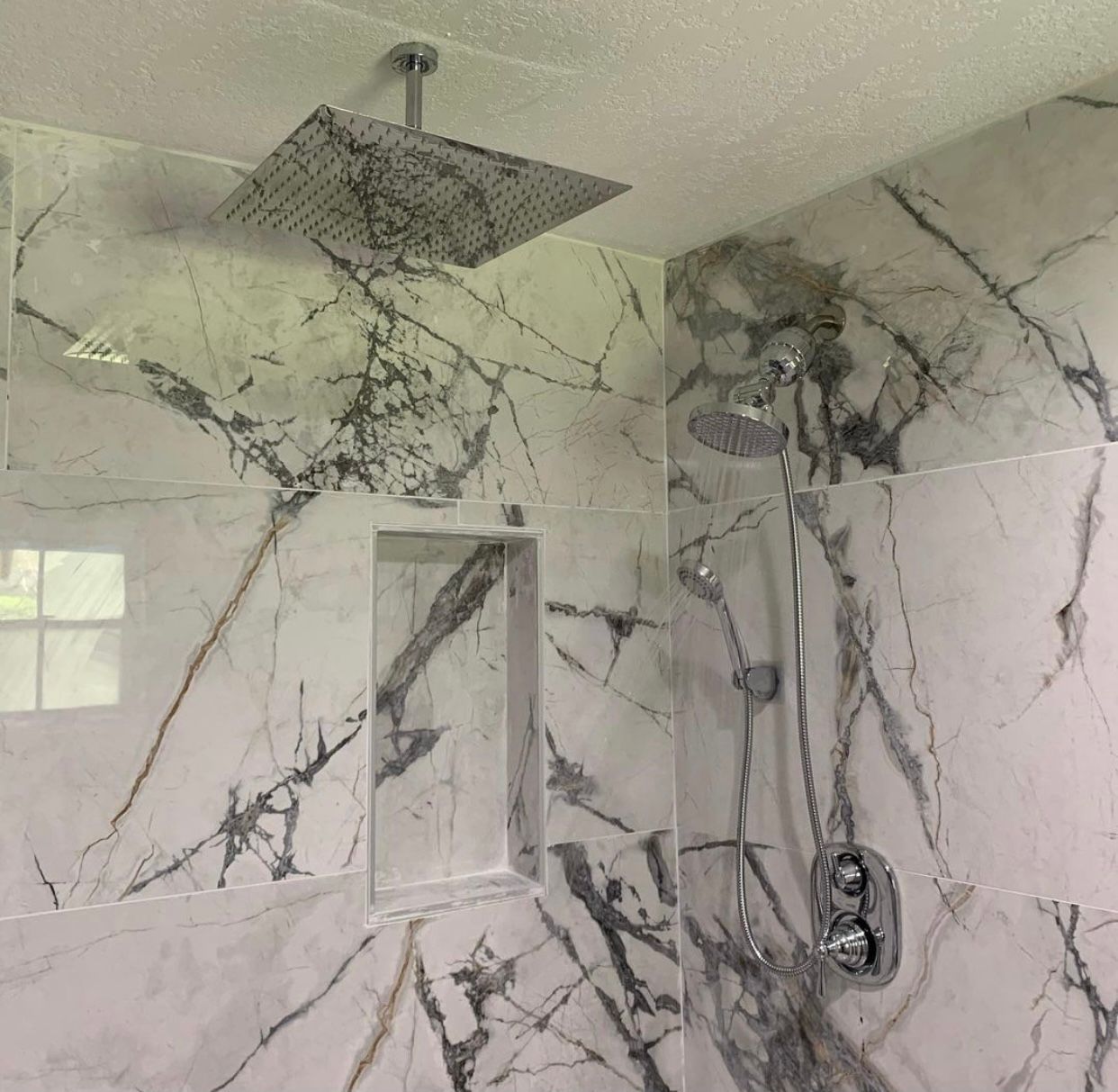 A modern shower stall with white and grey marble-patterned tiles, a square rainfall showerhead, and a handheld nozzle.