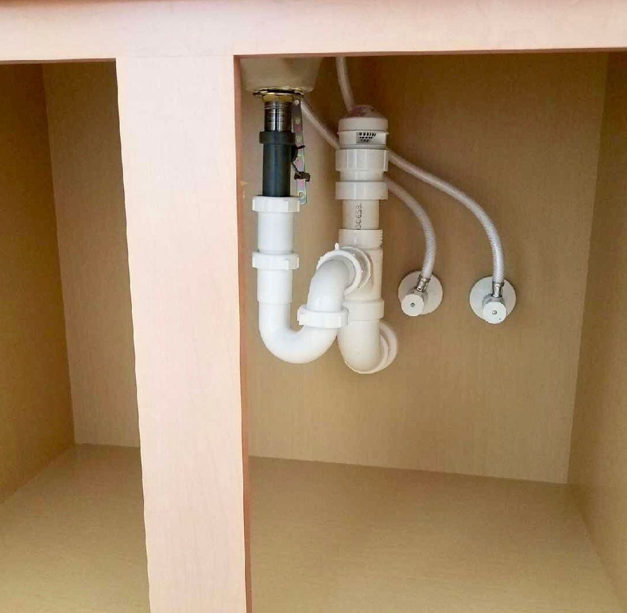 Under-sink plumbing showing a white PVC P-trap connected to drainage and water supply valves against a beige cabinet.