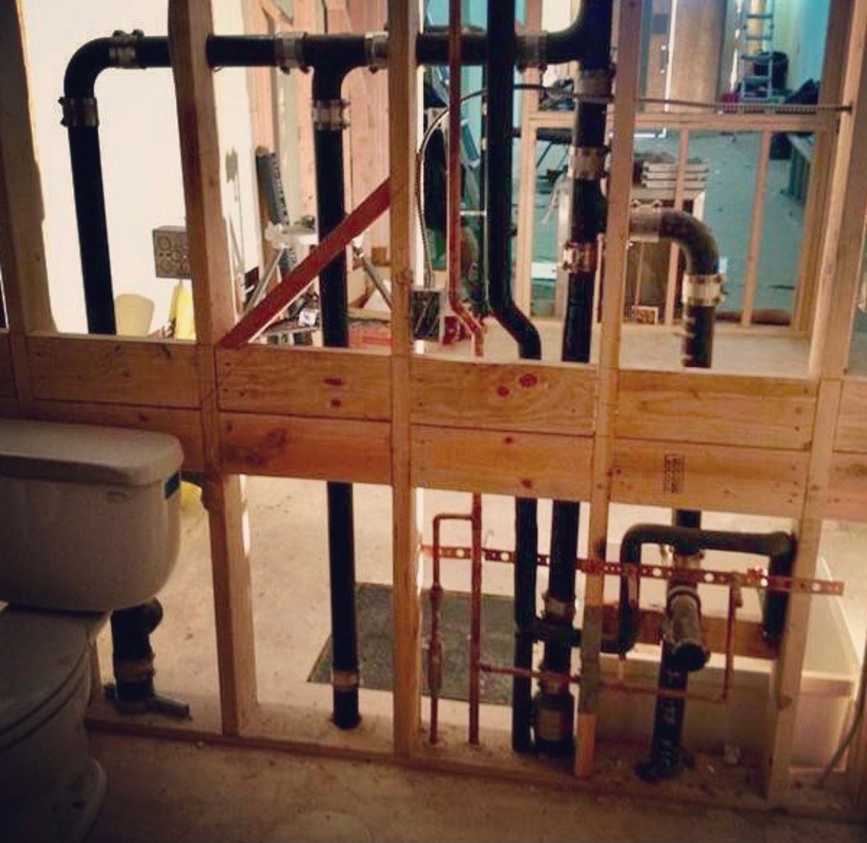 Exposed wall framing showing black ABS plumbing pipes and copper water supply lines behind a toilet in a room.