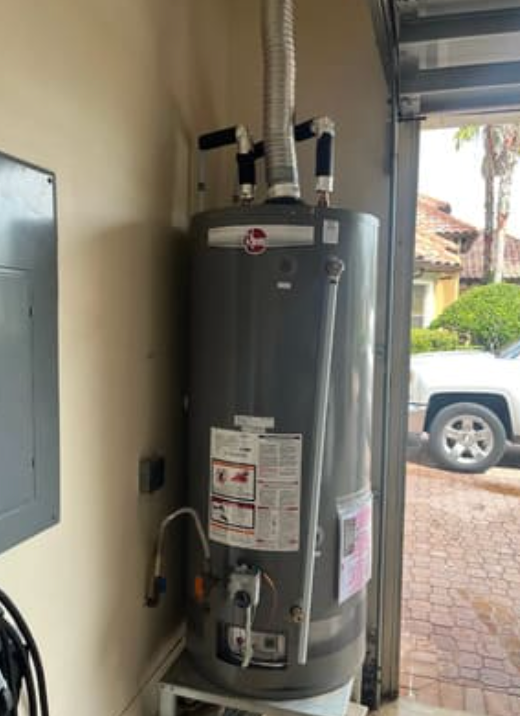 A gray Rheem water heater standing on a platform in a garage, next to an electrical panel and an open doorway.