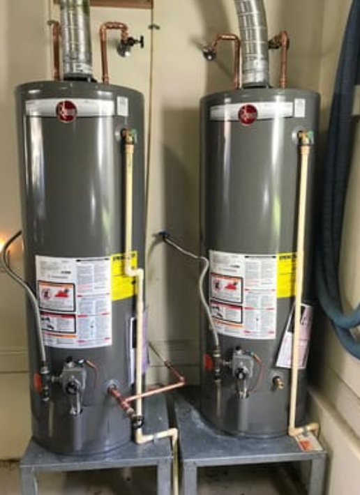 Two tall, gray Rheem gas water heaters on metal stands, connected by copper piping and vent ducts in a utility room.