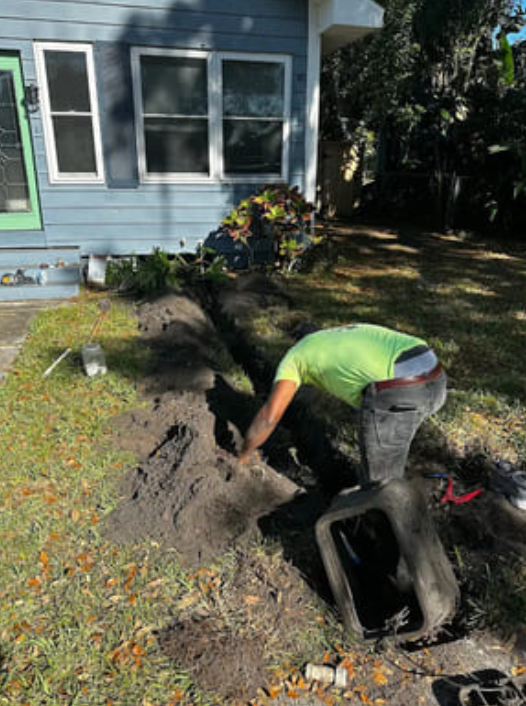 A person in a neon shirt digs a trench in a grassy front yard next to a light blue house.