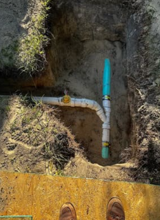 A dug-out trench showing white and blue PVC pipes connected in a T-junction with a yellow valve handle in the ground.