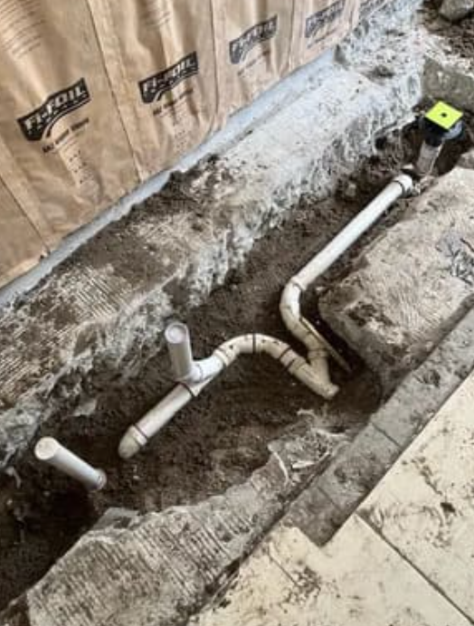 A plumbing system of white PVC pipes installed in a trench next to an insulated wall.