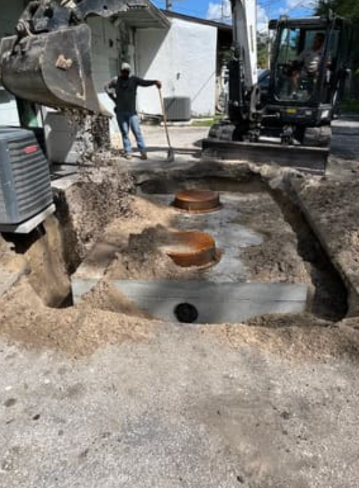 An excavator and worker at a construction site exposing a concrete septic tank with two access lids in the ground.